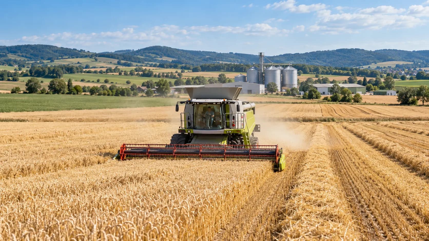 A combine harvester cutting a wheat field, with grain silos and farm buildings on the far side